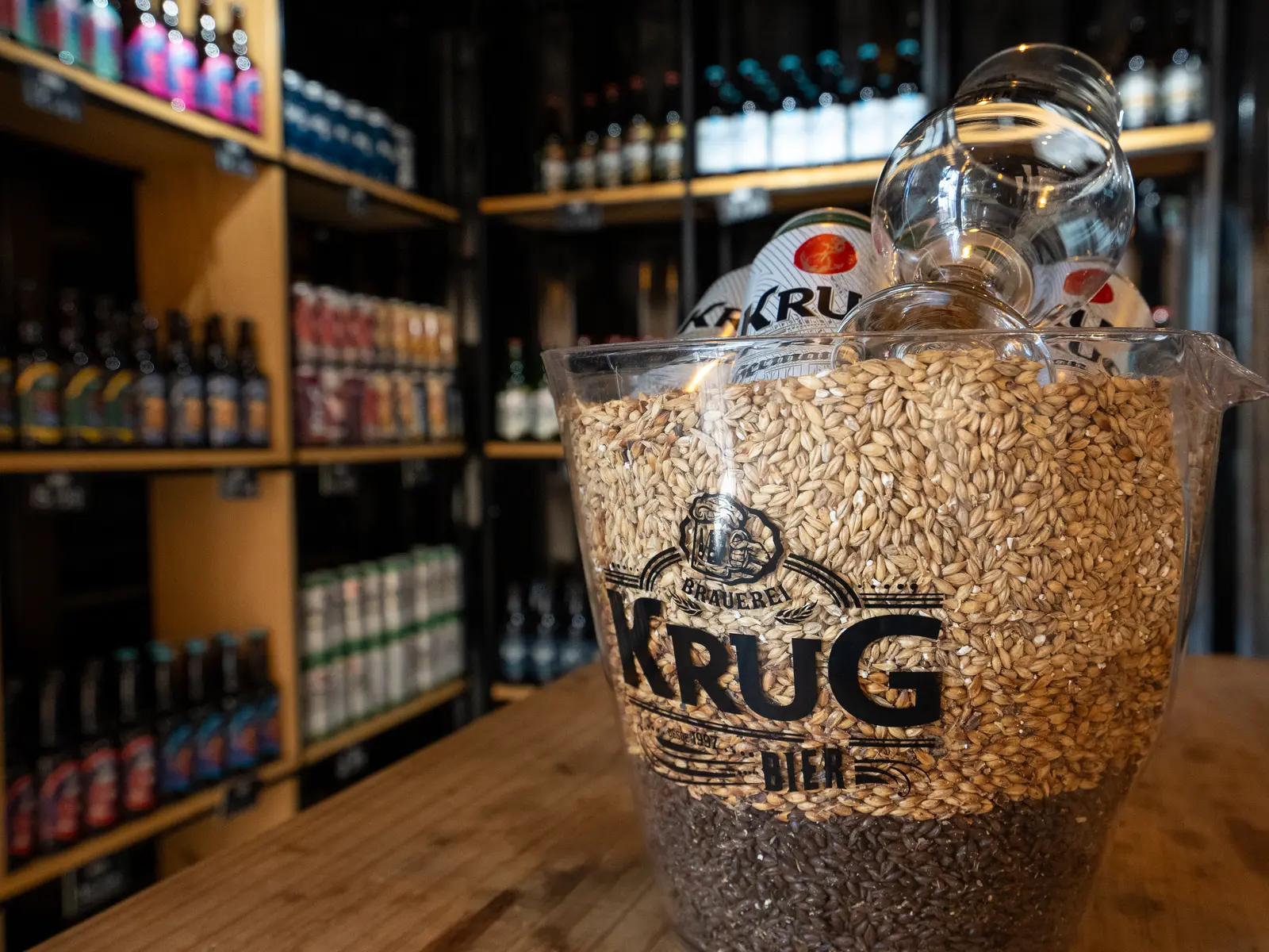 Balde de vidro cheio de malte cervejeiro sobre uma mesa de madeira na loja da fábrica da Krug Bier, com prateleiras de cervejas ao fundo