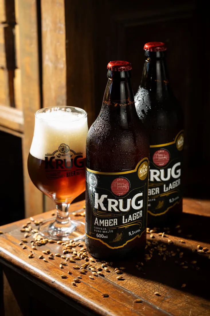 Taça de cerveja Krug Amber Lager ao lado de garrafas, com grãos de malte sobre a mesa
