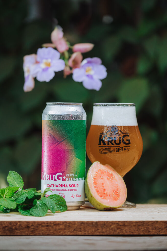 Lata e taça da Catharina Sour de goiaba e hortelã da Krug com a Cerveja Blumenau, com goiaba cortada e hortelã sobre a mesa