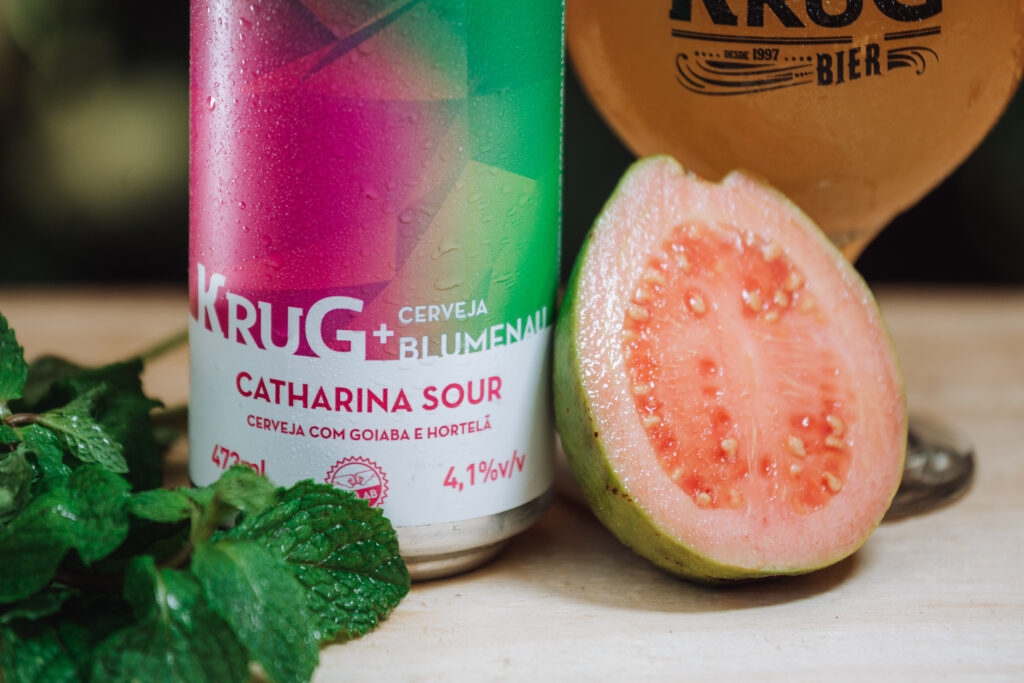 Lata da Catharina Sour de goiaba e hortelã da Krug com a Cerveja Blumenau, ao lado de uma goiaba cortada e folhas de hortelã