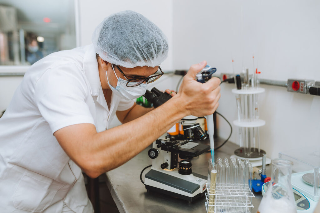 Profissional de jaleco e touca analisando uma amostra no microscópio no laboratório da fábrica da Krug Bier