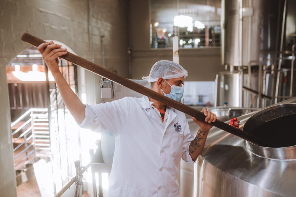 Cervejeiro de jaleco branco e touca movimentando o mosto com uma pá de madeira dentro da panela de brassagem na fábrica da Krug Bier